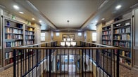 This two-story space is thoughtfully designed to foster comfort and community. At its heart is an open mezzanine level overlooking the floor below, framed by an elegant wrought-iron railing with a warm wood cap rail. A chandelier hangs gracefully above the open atrium, casting soft light across both levels and creating a welcoming focal point.

The upper level is lined with generously stocked built-in bookshelves, forming an inviting library that wraps around the open balcony. Patterned carpeting in rich tones of gold and navy runs throughout the hallways and common areas, adding warmth and visual depth. The corridors extend with ceiling fans overhead and recessed lighting, maintaining a well-lit and comfortable atmosphere.

A dedicated computer lounge features rich, dark wood cabinetry and desks equipped with workstations, complemented by patterned carpeting and ceiling-mounted lighting fixtures that give the room a studious yet cozy feel. Nearby seating nooks with accent chairs and artwork-adorned walls offer quiet retreats for relaxation or leisure activities such as board games.

Natural light filters in through multiple windows on both levels, brightening the common areas. Crown molding, tray ceiling details with wood trim accents, and framed artwork throughout contribute to the refined, residential character of this well-appointed multi-level community space.