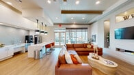 Located at 691 Union Chapel Rd in Cedar Creek, TX, this single-floor community space offers a thoughtfully designed open-plan layout that seamlessly blends a fully equipped kitchen with a spacious living area. The kitchen is outfitted with stainless steel appliances, including a French door refrigerator and a commercial-grade coffee station, all complemented by crisp white cabinetry, a textured tile backsplash, and a large white quartz island with bold orange velvet bar seating. Striking pendant light fixtures with brushed metal and white shades add a contemporary flair overhead. The living area features warm honey-toned hardwood floors throughout, a generous cognac leather sectional sofa, and a built-in wall niche housing a mounted flat-screen TV and linear fireplace, flanked by decorative alcoves with floral accents. Soaring ceilings with tray detailing and wood-paneled insets elevate the architectural character of the space. Large floor-to-ceiling windows and sliding glass doors flood the interior with natural light and provide views of the surrounding outdoor amenity areas. A dedicated work area includes a built-in desk station with dual monitors, set against a shimmering accent wall. Additional seating nooks throughout create versatile, inviting zones for relaxation or casual gatherings.