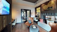 Located in Flower Mound, this single-floor community space blends modern design with warm, rustic elements to create an inviting and functional environment. The lounge area is anchored by rich, wide-plank hardwood flooring and striking reclaimed wood accent walls that add texture and depth throughout. A built-in business center workstation, framed by dark-trimmed glass partitions and pendant lighting, provides a dedicated computer workspace complete with an ergonomic chair — ideal for residents needing a quiet area to work or study. The seating area features a curved cognac leather swivel chair, a plush sofa with coordinating throw pillows, and a woven ottoman serving as a coffee table, creating an approachable and stylish gathering space. Large black-framed windows flood the interior with natural light while offering direct views of an outdoor patio furnished with lounge seating and shaded umbrellas. The elevated ceiling with a dark painted tray detail adds architectural interest and a sense of volume to the space. Double glass-paned entry doors with dark metal frames provide a sophisticated entry point, while pendant and recessed lighting ensure a warm, well-lit ambiance throughout the day and evening. The overall design strikes a cohesive balance between industrial and contemporary elements.