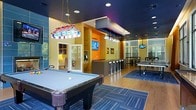 This dynamic single-floor amenity lounge is designed to serve as a central hub for recreation and socializing. The space features a bold, contemporary aesthetic defined by a striking dark ceiling that contrasts with warm wood-toned accent walls and vivid orange highlights, creating an energetic and inviting atmosphere throughout.

At the forefront of the room, a full-sized pool table takes center stage, illuminated by a decorative stained-glass pendant light that adds a classic gaming parlor feel. Adjacent to this area, a sleek bar counter with modern bar stools and pendant lighting provides a casual gathering spot, complemented by a mounted flat-screen TV for entertainment. An additional TV is positioned in a secondary lounge zone, ensuring visibility from multiple areas.

Beyond the bar, the space opens into a dedicated area for table tennis, set atop area rugs over hardwood-style flooring that zones the room effectively. Large windows along one wall allow natural light to filter in, adding brightness and a connection to the outdoors. A quieter seating nook is also visible, offering a relaxed retreat within the otherwise lively environment. The overall layout is open and well-organized, balancing activity zones with social spaces.