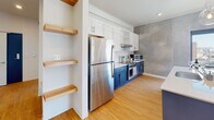 This single-floor unit showcases a thoughtfully designed contemporary interior that blends industrial and modern aesthetics throughout. The open-concept living and kitchen area serves as the centerpiece of the space, anchored by a striking kitchen with bold navy-blue lower cabinetry contrasted by crisp white upper cabinets. A large island with a white countertop and pendant globe lighting adds both functionality and visual interest. Stainless steel appliances, including a full-size refrigerator and range, complete the kitchen. Open wood shelving near the kitchen entry provides additional storage and display space.

Textured concrete-finish accent walls appear throughout the unit, lending an industrial character that complements the warm wood-look flooring running seamlessly across all main areas. Large windows and sliding glass doors flood the living area with natural light and provide outdoor views. A dedicated entryway features built-in wood cabinetry overhead and a bench, offering a practical transition zone.

The bedrooms are well-proportioned, each featuring the concrete accent wall, a window with a roller shade, and baseboard heating. The bathroom is generously sized and fully accessibility-equipped, with grab bars, a wood-toned vanity, tile flooring, and a curtained shower area. Recessed lighting throughout maintains a clean, modern ceiling profile.