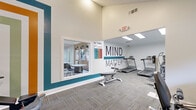 Located at 3600 Park Colony Dr in Norcross, Georgia, this single-floor fitness center is thoughtfully designed to support a well-rounded workout experience. The space is anchored by a bold "MIND MATTER" wall mural in teal and white, serving as a motivational focal point throughout the facility. The main workout area features a diverse selection of equipment, including treadmills, an elliptical trainer, a recumbent bike, a multi-station cable machine by Life Fitness, and adjustable weight benches, catering to both cardio and strength training needs. Floor-to-ceiling mirrors along one wall visually expand the space, while large windows invite natural light and offer views of the surrounding greenery. The flooring throughout consists of durable, multi-toned carpet tiles that provide comfort and practicality. Colorful horizontal stripe accents on the walls add a lively, energetic aesthetic to the strength training zone. An adjacent room, visible through a pass-through window, provides a quieter lounge or meeting area with seating and wall-mounted décor. A dedicated entry and exit point with glass doors connects the fitness center directly to the outdoors. A Body Mass Index chart and hand sanitizer station reflect attention to health-conscious amenities. The overall ambiance is vibrant, clean, and welcoming.
