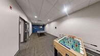 This recreational facility spans a generously sized single floor, thoughtfully designed to accommodate a wide variety of games and leisure activities. The space is divided into two interconnected areas, each with its own distinct character and function.

The main lounge and gaming area features bold red accent walls, two pool tables, an air hockey table, and multiple wall-mounted flat-screen TVs, creating a lively and energetic atmosphere. Comfortable gray sofas provide a relaxed seating area for spectators or those taking a break between games. Exposed industrial-style ductwork runs along the ceiling, adding a modern edge to the overall aesthetic, while recessed lighting and a drop-panel ceiling ensure the space is well-lit throughout.

The secondary area offers a dedicated ping pong room and foosball table, set against a deep blue accent wall adorned with decorative shelving and wall art, lending a slightly more intimate feel. Both sections are finished with patterned, durable carpet tiles that complement the contemporary interior design.

The layout flows naturally between the two zones through an open doorway, making the entire space feel cohesive and accessible. Together, these rooms form a dynamic, multi-use recreational environment well-suited for community gatherings, team-building activities, or casual everyday use.