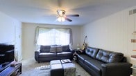 This single-floor residence offers a welcoming and functional layout designed for comfortable everyday living. The living room serves as the heart of the space, featuring a large window dressed with sheer draping curtains that allow generous natural light to fill the room, creating an airy and inviting atmosphere. The ceiling is finished with a classic textured treatment and outfitted with a multi-blade ceiling fan with integrated lighting, providing both air circulation and ambient illumination. Soft carpeted flooring runs throughout the living area, adding warmth underfoot. The room comfortably accommodates generous seating with two dark leather sofas — one a classic sofa with accent throw pillows, the other a tufted sectional-style piece — arranged around a central ottoman set that doubles as additional surface space. The neutral-toned walls keep the space feeling open and versatile. Overall, the living area strikes a balance between practicality and comfort, offering a relaxed residential environment well-suited for both quiet evenings and casual gatherings.