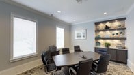 This single-floor professional meeting space is thoughtfully designed to support productive gatherings and formal discussions. The conference room serves as the centerpiece of the space, featuring a large oval dark-wood meeting table surrounded by high-back black leather executive chairs, providing comfortable seating for multiple attendees. Recessed lighting in the white ceiling, accented by elegant crown molding, ensures a well-lit and polished environment.

Natural light filters generously into the room through multiple windows fitted with white horizontal blinds, creating a bright yet controlled atmosphere. A standout architectural feature is the built-in stone-clad accent wall with illuminated open shelving, adding texture and a refined aesthetic to the space. Decorative accessories in teal and blue tones are displayed on the shelves, complementing the room's cool gray and neutral color palette.

A credenza positioned near the windows supports office equipment, adding functional utility without disrupting the room's professional aesthetic. A framed landscape artwork hangs on the wall, contributing to the calm and focused ambiance. A patterned area rug anchors the seating arrangement, adding warmth to the overall design. This space is well-suited for meetings, presentations, and professional gatherings.
