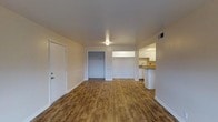 This single-floor unit offers a straightforward, functional layout designed for comfortable everyday living. The main living area is finished with warm-toned hardwood-style flooring that runs throughout, lending a cohesive and inviting feel to the space. Neutral white walls and a clean, minimalist aesthetic allow for versatile interior styling and decoration.

A notable feature of the living area is a built-in closet with a dedicated hanging rod, providing convenient built-in storage directly off the main room. The front entry door opens directly into this bright, open space, creating an accessible and welcoming arrival point.

The kitchen is situated in an open nook adjacent to the living area, allowing for an easy flow between spaces. It is equipped with white upper cabinetry, a countertop with a sink, and lower cabinet storage, offering a practical and tidy cooking environment. A HVAC vent is visible, indicating climate control throughout the unit.

Overhead lighting in the living area ensures the space remains well-lit regardless of natural light availability. The overall ambiance is clean, move-in ready, and well-suited for individuals or small households seeking a practical, no-frills living environment on a single level.