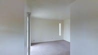 This single-story space presents a clean, minimalist interior defined by its open layout and neutral palette. The room visible is finished with wall-to-wall carpet in a muted taupe tone, providing a soft, comfortable underfoot surface that contributes to the room's understated aesthetic. White walls are kept simple and unadorned, offering a blank canvas adaptable to a variety of uses and furnishing styles.

The textured ceiling adds subtle visual interest while maintaining the room's overall light and airy feel. A narrow, vertically oriented window positioned along the far wall serves as a key architectural detail, drawing natural light deep into the interior and creating a bright, welcoming atmosphere despite its slender profile. An additional light source is visible from a doorway or opening to the left, suggesting the space connects to other areas that further contribute to the overall brightness.

The room's proportions appear comfortable and functional, with clean lines throughout. Electrical outlets are conveniently placed along the baseboards. The overall ambiance is calm and neutral, making this space well-suited for customization to meet a range of living or functional needs.