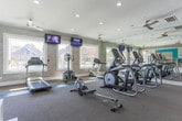 Located at 101 Peaks Landing in Acworth, Georgia, this well-appointed fitness center offers a comprehensive workout environment designed for a variety of exercise routines. The space is thoughtfully laid out across a single floor, featuring distinct zones for cardio training, strength work, and cycling. A signature teal color scheme runs throughout, accenting the neutral gray walls, carpeted flooring, and structural columns to create a cohesive, energetic atmosphere.

The main cardio area is generously proportioned, housing a lineup of treadmills, ellipticals, and a rowing machine, all positioned to take advantage of large windows that let in abundant natural light and offer views of the surrounding outdoor amenity area, including a pool. Wall-mounted flat-screen televisions are positioned throughout for entertainment during workouts. Ceiling fans provide additional airflow and comfort.

A dedicated cycling studio features a row of stationary spin bikes, each paired with its own personal mounted screen for an immersive ride experience. Teal accent panels frame the screens, reinforcing the facility's bold design identity.

The free weight and functional training area includes a full dumbbell rack, open floor space, and built-in shelving for equipment storage. Full-length mirrors expand the sense of space and support proper form. Crown molding and recessed lighting add a polished, refined finish to every corner of this impressive fitness facility.