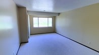 This single-floor space presents a clean, functional layout centered around a generously sized main room. The room is finished with wall-to-wall carpeting in a neutral gray-blue tone, providing a soft, comfortable underfoot feel throughout the space. The walls are painted in a warm beige, creating a understated and versatile backdrop suitable for a variety of uses.

A prominent multi-pane window dominates one wall, allowing natural light to filter into the room while framing a pleasant view of surrounding greenery and trees. This window serves as a key architectural feature, contributing to an open and airy atmosphere despite the room's straightforward design.

The ceiling features a classic textured finish, adding subtle visual interest overhead. A centrally placed air vent is visible, indicating a climate control system integrated into the structure. Dark baseboards provide a defined contrast against both the carpeted floor and the lighter walls, lending the room a clean, finished appearance.

A doorway is visible along one side, suggesting additional rooms or storage areas connected to this main space. Overall, the space offers a bright, neutral environment with practical finishes and a calm, uncluttered ambiance.