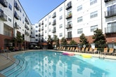 This outdoor amenity space is a well-appointed retreat designed for relaxation and leisure. At its centerpiece is a generously sized, crystal-clear swimming pool with easy-access entry rails, perfect for unwinding on warm days. The surrounding pool deck is finished in smooth concrete and offers ample space for lounging, with multiple sling-style chaise lounge chairs accented by yellow headrest cushions and paired with slatted side tables for convenience. Vibrant orange market umbrellas provide shaded seating areas with accompanying patio furniture, ideal for gathering or casual dining outdoors. The space is framed by mature magnolia trees, manicured shrubs, and lush greenery that add a natural, park-like ambiance to the urban setting. The community building itself serves as a striking architectural backdrop, featuring a bold mix of brick, white paneling, and vivid teal and orange accent walls that lend a modern, energetic character to the environment. Upper-level balconies overlook the courtyard, reinforcing the sense of an active, connected community. The entire outdoor area is enclosed by decorative iron fencing, ensuring privacy and security while maintaining an open, airy feel throughout this thoughtfully designed communal space.