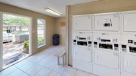 Located at 5440 N Jim Miller Rd in Dallas, Texas, this single-story shared laundry facility is designed to serve residents with a practical and functional layout. The room features light-toned tile flooring throughout, providing a clean and easy-to-maintain surface. Warm beige walls with a wainscoting detail lend a simple, tidy aesthetic to the space.

A row of top-load washing machines lines one wall, each equipped with card-operated payment systems for added convenience. Instructional signage for both washing and drying is posted prominently above the machines, offering clear guidance for users. On the opposite end of the room, stacked dryer units provide ample drying capacity, also featuring card-based payment terminals.

A folding table offers a practical surface for handling laundry, while benches provide seating while waiting. A bulletin board is mounted on the wall, serving as a community communication point. A fire extinguisher is visibly installed, ensuring safety compliance.

Natural light filters into the space through a window and a glass-paneled door, which opens to an outdoor area with surrounding greenery. Overhead fluorescent lighting ensures the room remains well-lit at all times. The space is completed with a waste bin for easy disposal, making this laundry facility a well-equipped and user-friendly amenity.