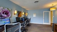 This single-floor business center offers a functional and comfortable environment designed to support productivity. The room is finished with soft blue walls that create a calm, focused atmosphere, complemented by warm wall sconce lighting and colorful botanical artwork that adds a touch of personality to the space. The centerpiece of the room is a long, dark-finished shared desk that accommodates multiple workstations, each equipped with desktop computers and ergonomic mesh chairs, allowing several users to work simultaneously. A shared printer is conveniently positioned along the desk, making document handling straightforward and efficient. The flooring features neutral-toned carpet that contributes to a quiet working environment, while a textured ceiling with a central ventilation vent ensures comfortable air circulation throughout. A framed informational notice is posted on the wall, suggesting this space is part of a larger managed facility. A door on the far side of the room provides easy access to adjacent areas. Overall, the business center balances practicality with a welcoming aesthetic, making it a convenient and pleasant resource for those needing access to technology and office amenities.