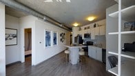 This 1-bedroom, 1-bathroom residence offers an open-concept layout that seamlessly blends industrial character with modern comfort. Exposed concrete ceilings and visible ductwork lend an urban, loft-inspired aesthetic, while wide-plank hardwood-style flooring grounds the space with warmth and continuity throughout.

The kitchen is well-appointed with white shaker cabinetry, black appliances including a range and refrigerator, a stainless steel microwave, and a sink with ample counter space. A central kitchen island with bar seating creates a natural gathering point and provides additional workspace, making it ideal for both cooking and casual dining.

The adjoining living area benefits from the open floor plan, creating a sense of spaciousness and easy flow between spaces. White walls contrast with the raw industrial ceiling elements, offering a clean, contemporary backdrop accented by thoughtful wall decor. A hallway leads to the private bedroom and bathroom, ensuring a comfortable separation between living and sleeping areas.

The single-floor layout makes navigation intuitive, and the overall design strikes a balance between urban industrial style and everyday functionality—making it a well-rounded and characterful living space.