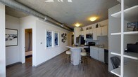 This 1-bedroom, 1-bathroom residence offers an open-concept layout that seamlessly blends industrial character with modern comfort. Exposed concrete ceilings and visible ductwork lend an urban, loft-inspired aesthetic, while wide-plank hardwood-style flooring grounds the space with warmth and continuity throughout.

The kitchen is well-appointed with white shaker cabinetry, black appliances including a range and refrigerator, a stainless steel microwave, and a sink with ample counter space. A central kitchen island with bar seating creates a natural gathering point and provides additional workspace, making it ideal for both cooking and casual dining.

The adjoining living area benefits from the open floor plan, creating a sense of spaciousness and easy flow between spaces. White walls contrast with the raw industrial ceiling elements, offering a clean, contemporary backdrop accented by thoughtful wall decor. A hallway leads to the private bedroom and bathroom, ensuring a comfortable separation between living and sleeping areas.

The single-floor layout makes navigation intuitive, and the overall design strikes a balance between urban industrial style and everyday functionality—making it a well-rounded and characterful living space.