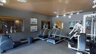 This fitness center offers a practical and inviting space designed to support a well-rounded workout routine. The room is finished with neutral grey walls and warm-toned ceilings, creating a focused yet comfortable atmosphere for exercise. Track lighting is mounted overhead, ensuring the space is well-lit throughout. The cardio area is a highlight of the room, featuring multiple treadmills positioned side by side, providing ample room for users to exercise simultaneously. A rowing machine is also available, rounding out the cardio offerings. On the opposite side of the room, a multi-functional strength training machine is available for resistance and weight-based workouts. The flooring consists of durable carpet suitable for a fitness environment. Windows with slatted blinds along the wall allow for natural light while maintaining privacy. Framed artwork adorns the walls, adding a polished, welcoming touch to the space. A wall-mounted clock ensures users can manage their workout time effectively. Overall, this fitness center is thoughtfully arranged to maximize functionality while maintaining an organized and aesthetically pleasing environment.
