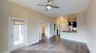 This single-story space showcases a contemporary open-concept layout that effectively connects the main living area with the kitchen. The living space features warm-toned hardwood-style flooring that extends throughout, complemented by tall ceilings that enhance the sense of openness and airiness. A ceiling fan with a built-in light fixture serves as a functional centerpiece overhead, improving both comfort and illumination.

The kitchen is positioned toward one end of the open floor plan and is equipped with stainless steel appliances, including a full-size refrigerator. A kitchen island with pendant lighting above provides additional workspace and defines the boundary between the cooking and living zones. Dark cabinetry contrasts with light countertops, adding visual depth to the space.

Multiple interior doorways lead to additional rooms, suggesting a well-organized floor plan with clear separation between living and private spaces. Natural light enters through a sliding glass door on one side, brightening the main living area. The neutral wall tones and clean white trim throughout create a cohesive, modern aesthetic that feels both inviting and functional.