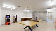 This single-floor recreational lounge is thoughtfully designed to serve as a dynamic social and leisure hub. The open-plan layout is anchored by a full-size JOOLA ping-pong table positioned at the center of the room, providing an immediate focal point and an inviting invitation to play. Light wood-look flooring runs throughout, contributing to the space's clean, contemporary aesthetic.

Along one wall, a sleek kitchenette features rich dark wood cabinetry with gold hardware and a white countertop with a built-in sink, offering a convenient refreshment station for guests. A playful wall mural reading "This Must Be The Place" adds a welcoming, artistic touch nearby. Two classic arcade machines — including a Pac-Man cabinet and a racing game — are positioned throughout the room, nodding to retro gaming culture.

Seating is distributed across multiple zones: high-top bistro tables with blush-toned bar stools create casual gathering spots, while a pair of olive-green lounge chairs arranged around a small side table offer a more relaxed seating nook. Glass-panel partitions and doors define the perimeter of the space, maintaining an open, airy feel while allowing visual connectivity. Overhead, recessed and fluorescent lighting ensures the room is well-lit throughout.