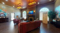 This single-story community clubhouse lounge is designed to provide a comfortable and functional gathering space with a warm, inviting ambiance. The main lounge area is immediately distinguished by its soaring ceilings, accented by a rich terracotta-toned upper wall and elegant crown molding. Two statement chandelier-style pendant light fixtures, complemented by recessed lighting and wall sconces, create a well-lit yet cozy atmosphere throughout.

The focal point of the lounge is a classic brick fireplace topped with a mounted flat-screen television, flanked by tall arched windows that flood the space with natural light and offer views of the surrounding greenery. The flooring throughout is a rich, dark wood-look tile laid in a striking herringbone pattern, adding a sophisticated design element to the space.

Seating is arranged thoughtfully, with plush red sofas and accent chairs providing ample gathering areas. Patterned accent chairs add visual interest near the fireplace. The entry area features an arched decorative niche with a gilded mirror above a built-in cabinetry unit, vivid abstract artwork, and additional seating. Adjacent rooms, accessible through French doors and arched doorways, appear to include an office or business center and additional amenity spaces.