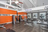 The Fieldcrest Fitness Center at 31 Trillium Cir in Dothan, Alabama is a well-appointed, single-room exercise facility designed to support a comprehensive workout experience. The space is defined by its striking vaulted ceiling with exposed white structural beams, which lend an open, airy feel and generous vertical height throughout. Hardwood-style flooring runs across the entire room, providing a clean and durable surface for all fitness activities.

The interior is boldly divided into two functional zones, clearly identified by large wall signage: a Strength area and a Cardio area. The accent walls are painted a vivid orange with white horizontal striping, creating an energizing atmosphere. The Strength zone is equipped with a cable machine, weight bench, dumbbell rack with a full range of free weights, medicine balls, resistance equipment, and a multi-functional cable tower. The Cardio zone features treadmills, elliptical trainers, a stationary bike, and a stair climber.

Natural light enters through multiple windows, offering views of the surrounding community and keeping the space bright and welcoming. Ceiling fans with integrated light fixtures provide additional ventilation and illumination. The overall design balances motivating aesthetics with practical functionality, making this fitness center a well-equipped destination for residents seeking a convenient and effective place to exercise.