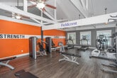 The Fieldcrest Fitness Center at 31 Trillium Cir in Dothan, Alabama is a well-appointed, single-room exercise facility designed to support a comprehensive workout experience. The space is defined by its striking vaulted ceiling with exposed white structural beams, which lend an open, airy feel and generous vertical height throughout. Hardwood-style flooring runs across the entire room, providing a clean and durable surface for all fitness activities.

The interior is boldly divided into two functional zones, clearly identified by large wall signage: a Strength area and a Cardio area. The accent walls are painted a vivid orange with white horizontal striping, creating an energizing atmosphere. The Strength zone is equipped with a cable machine, weight bench, dumbbell rack with a full range of free weights, medicine balls, resistance equipment, and a multi-functional cable tower. The Cardio zone features treadmills, elliptical trainers, a stationary bike, and a stair climber.

Natural light enters through multiple windows, offering views of the surrounding community and keeping the space bright and welcoming. Ceiling fans with integrated light fixtures provide additional ventilation and illumination. The overall design balances motivating aesthetics with practical functionality, making this fitness center a well-equipped destination for residents seeking a convenient and effective place to exercise.