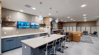 This well-appointed community space combines a fully equipped kitchen with a comfortable living room, all contained on a single floor. The kitchen area is anchored by a large central island topped with a white marble-style countertop, surrounded by sleek black bar stools with chrome accents. Globe-shaped pendant lights hang overhead, adding a modern touch. Navy blue cabinetry lines the walls, complemented by light-colored tile backsplash and marble countertops. Multiple wall-mounted flat-screen TVs are positioned above the cabinetry, making the space ideal for group entertainment. A long bar-height counter along one wall provides additional seating and workspace. The living room area transitions seamlessly from the kitchen, featuring a tan leather sofa, black accent chairs, and a low coffee table set atop a patterned area rug. A large TV and a built-in wooden bookcase with generous shelving for books and décor serve as a focal point in this area. Throughout the space, the walls are finished with a warm textured wallpaper above gray wainscoting panels, creating a cohesive and refined aesthetic. Light-toned tile flooring, recessed lighting, and a neutral color palette tie the entire space together into a functional and inviting communal environment.