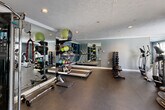This fitness center offers a thoughtfully designed single-floor workout space suited for a wide range of exercise routines. The room features a two-tone color scheme, with deep navy accent walls contrasting against light gray surrounds, giving the space a clean, modern aesthetic. Crown molding and recessed lighting add a polished finish, while a textured ceiling completes the interior detail.

Floor-to-ceiling mirrors line one wall, visually expanding the space and allowing users to monitor their form during workouts. The durable dark rubber flooring provides excellent support and is well-suited for high-impact activity throughout the room.

Cardio equipment is well-represented, with multiple treadmills, an elliptical trainer, a stationary bike, and a balance board available for use. Strength training options are equally comprehensive, including a multi-functional cable machine, a full rack of dumbbells, kettlebells, medicine balls, resistance ropes, and various weighted accessories organized on dedicated storage racks.

Natural light filters in through several windows fitted with white shuttered blinds, particularly in the cardio area, where outdoor greenery is visible. A glass-paneled entry door adds to the sense of openness. The space also includes a towel rack, a waste bin, and wall-mounted informational signage, ensuring a fully functional and user-friendly environment.