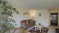This single-story space presents a straightforward and functional layout centered around a welcoming living area. The living room features neutral white walls and light-toned flooring, creating a clean and airy backdrop throughout. A plush, tan microfiber sofa serves as the focal point of the seating area, accented with colorful throw pillows in warm orange and floral patterns. A bold floral-patterned area rug in dark tones anchors the space, adding visual contrast against the lighter flooring. A small white side table complements the seating arrangement, providing a practical surface for everyday use. Soft ambient lighting is provided by a floor lamp positioned in the corner, contributing to the room's relaxed atmosphere. Decorative wall accents, including floral wall art and a charming birdcage ornament, add personality without overwhelming the space. Lush green houseplants placed near the walls bring a touch of nature indoors, enhancing the overall warmth of the interior. The space maintains a modest and comfortable scale, with clean lines and simple décor choices that create an approachable, lived-in feel throughout.