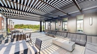 Located at 110 Woodcrest Rd in Cherry Hill, NJ, this sophisticated outdoor terrace is designed to deliver a premium open-air experience. The space is defined by a striking dark-framed pergola that spans the entire deck, casting dramatic linear shadows across the large-format tiled flooring and creating a visually dynamic atmosphere throughout the day. Transparent glass railings line the perimeter, preserving unobstructed panoramic views of the surrounding residential community and lush greenery beyond. The terrace is organized into distinct functional zones. A generous lounge area features a continuous row of modern, upholstered seating arranged along a sleek dark panel wall with integrated wall sconces, centered around a contemporary fire table — ideal for relaxed gatherings. Multiple outdoor dining tables with coordinating chairs are positioned throughout the open deck, accommodating a variety of group sizes. A central access point with full-glass double doors connects the terrace to the adjacent interior, while a metal-and-glass staircase provides additional access from ground level. The combination of the architectural pergola, clean material palette of dark metal, glass, and neutral stone tile, and the abundant natural light flooding the space creates a refined, resort-style outdoor amenity.