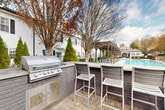 Located at 575 McDonough Pkwy in McDonough, Georgia, this outdoor community amenity space is designed to provide residents with a well-appointed and functional gathering environment. The centerpiece of the space is a built-in outdoor kitchen featuring a stainless steel multi-burner gas grill set into a brick-constructed surround, complemented by an extended bar-height counter with modern slatted bar seating — ideal for casual outdoor dining and entertaining.

Adjacent to the grilling area, a covered pergola-style pavilion with string lighting overhead offers a shaded lounge space, furnished with comfortable seating, providing a welcoming retreat in any weather. The open patio area is finished with a combination of concrete and paver surfaces, creating a clean and durable outdoor floor plan.

A sparkling swimming pool is visible nearby, lined with lounge chairs and framed by a pergola structure, adding a resort-like quality to the amenity area. The surrounding grounds are thoughtfully landscaped with mature trees, evergreen shrubs, ornamental grasses, and seasonal plantings, all enclosed by a black iron perimeter fence that provides both security and aesthetic definition. Concrete walkways connect the various zones of the space, ensuring easy accessibility throughout.