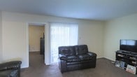 Located at 160 Broadway St in Clinton, Mississippi, this single-floor residence offers a straightforward and functional layout designed for comfortable everyday living. The living area serves as the central gathering space, characterized by neutral-toned walls and wall-to-wall carpeting that adds warmth underfoot. A large sliding glass door dressed with vertical blinds allows generous natural light to fill the room while providing access to the outdoors. The open feel of the space is enhanced by clean, smooth ceilings and a simple, uncluttered design. An adjoining room is visible through an interior doorway, suggesting a connected and practical floor plan that allows for easy movement throughout the home. The overall ambiance is calm and understated, making it a versatile space suited to a variety of furnishing styles and uses. The single-story layout offers convenience and accessibility throughout the home.