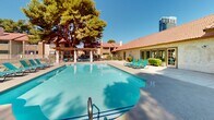 Located at 4201 W Rochelle Ave in Las Vegas, Nevada, this single-level outdoor amenity area is designed for relaxation and recreation in the heart of the desert Southwest. The centerpiece of the space is a generously sized, freeform swimming pool with clear blue water, surrounded by a spacious concrete pool deck outfitted with teal lounge chairs ideal for sunbathing under the abundant Nevada sunshine. Adjacent to the main pool is a separate spa and hot tub, nestled within a landscaped setting framed by mature pine and cypress trees that provide natural shade and a sense of privacy.

A substantial wood-and-masonry pergola structure anchors one side of the deck, offering a shaded outdoor lounge area furnished with teal seating and a coffee table — a welcoming retreat from the sun. The pergola's open slatted roof allows filtered light to pass through while maintaining a cool, comfortable environment beneath.

Overlooking the pool area is a single-story clubhouse building featuring a distinctive red clay tile roof, stucco exterior, and large French-style windows and doors that open directly onto the pool deck. Well-maintained greenery, shrubs, and landscaping border the perimeter, and a secured fence encloses the entire area. Views of the surrounding Las Vegas skyline are visible in the background, adding an urban backdrop to this resort-style outdoor setting.