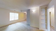 This single-floor space features an open living area designed with both functionality and comfort in mind. The main living space is carpeted and generously sized, with a ceiling fan centrally positioned to ensure airflow throughout. A large window fitted with horizontal blinds allows natural light to filter into the room, creating a bright and welcoming atmosphere.

A textured ceiling adds subtle visual interest overhead, while clean white walls and neutral tones give the space a versatile, move-in-ready feel. A decorative wall sconce provides additional ambient lighting along one wall, complementing the natural light from the window.

One of the standout features of this space is the built-in shelving unit positioned along one wall, offering organized storage and display options. A partial wall or column separates areas of the open floor plan, providing a sense of definition between spaces without enclosing them entirely. A secondary light fixture is visible beyond this dividing column, suggesting an additional functional zone within the layout. The overall ambiance is clean, neutral, and well-lit, making it adaptable to a variety of interior design preferences.