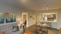 This single-floor residence showcases an open-concept layout that integrates the living, dining, and kitchen areas into one cohesive, functional space. The living area is appointed with soft, neutral-toned walls that create a warm and inviting atmosphere, complemented by dark hardwood-style flooring that runs throughout. A comfortable seating arrangement anchors the living space, accented by a stylish table lamp and contemporary wall art.

The dining area flows naturally from the living room, featuring a sleek dining table with upholstered chairs, positioned conveniently adjacent to the kitchen. The kitchen is visible in the background, highlighted by pendant lighting and stainless steel appliances, including a full-size refrigerator, and a light-toned countertop that adds contrast to the overall color palette.

A hallway visible from the main living area leads to a bathroom, offering a practical layout that maximizes accessibility. The space benefits from recessed lighting and a neutral color scheme throughout, contributing to a cohesive and modern aesthetic. The open floor plan and thoughtful design make this residence feel both spacious and efficiently organized.
