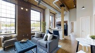 Located at 390 Capitol Ave, this single-floor loft showcases a compelling blend of historic industrial character and modern comfort. The space is defined by its dramatically tall ceilings, which feature exposed wooden planks, original timber beams, and large-diameter ductwork—hallmarks of a thoughtfully preserved industrial conversion. Structural wood columns with warm natural finishes anchor the open floor plan, adding architectural interest throughout.

Oversized, multi-pane factory-style windows dominate the exterior wall, drawing in abundant natural light and offering views of the surrounding neighborhood. The exposed brick walls provide rich texture and a sense of history, complementing the light-toned hardwood flooring that runs throughout the space.

The living area is generously proportioned, accommodating comfortable seating with room to spare. It transitions seamlessly into the kitchen, which features dark cabinetry, a breakfast bar peninsula, and full-size appliances—offering both functionality and an open connection to the main living space. Track lighting and ceiling fans complete the practical yet stylish overhead details. The overall ambiance is warm, airy, and distinctly urban.