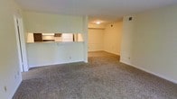 This single-floor residential space offers a practical and comfortable layout designed for everyday living. The open-concept living area is finished with wall-to-wall carpeting in a neutral tone, creating a warm and inviting atmosphere throughout. A defining architectural feature is the pass-through counter that connects the kitchen to the living space, promoting an open flow between the two areas while maintaining a sense of separation. The kitchen is equipped with dark wood cabinetry, a white refrigerator, and a gas range, providing all the essential amenities for meal preparation. The living area extends generously, offering ample room for furniture arrangement and day-to-day activities. The walls throughout are finished in a soft, neutral palette that enhances the brightness of the space. A built-in shelf or ledge detail is visible along one wall of the living area, adding a subtle architectural element. Overhead lighting is present, and HVAC vents are integrated into the ceiling, ensuring year-round comfort. White baseboards frame the carpeted floors cleanly throughout. Overall, the space presents a straightforward, well-maintained interior that prioritizes function and ease of living.