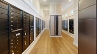 Located at 415 Curtis Pkwy SE in Calhoun, Georgia, this dedicated mail and package room serves as a centralized hub for resident correspondence and deliveries. The hallway-style space is thoughtfully designed with a clean, modern aesthetic, featuring warm wood-look flooring, crisp white walls with panel molding details, and recessed lighting that keeps the room bright and welcoming. Rows of numbered black metal mailboxes line both sides of the room, providing individual secure mail access for a large number of residents, with unit numbers clearly labeled on each door for easy identification. A standout feature of the room is the Package Concierge smart locker system, a sleek, dark-finished automated unit that allows residents to retrieve packages securely at their convenience. High transom windows near the ceiling allow natural light to filter in, enhancing the open feel of the space. The room is organized with a central structural column that divides the layout while maintaining clear sightlines throughout. White baseboard trim and panel detailing add a polished, residential quality to the utilitarian function of the space. Overall, the room combines practical functionality with a refined design, ensuring an efficient and pleasant mail retrieval experience for all residents.