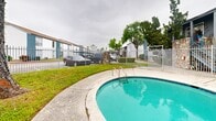 Located at 6500 W 43rd St in Houston, Texas, this single-level outdoor community space centers around a generously sized freeform pool with a distinctive kidney-shaped design. The pool's clear turquoise water is complemented by decorative tile detailing along the shallow entry, and a "No Diving" designation ensures safe use for all residents. Metal pool ladders provide convenient entry and exit points.

The surrounding concrete deck offers ample space for lounge seating and poolside activities. A secure wrought-iron perimeter fence with a gated entrance encloses the area, providing both safety and privacy. Stone-accented entry columns add a touch of architectural character to the gating.

Adjacent to the pool, a small utility structure with a blue metal roof serves as a functional amenity building. Mature trees are positioned throughout the grounds, offering natural shade and greenery. A grassy lawn area borders the pool deck, adding to the outdoor atmosphere.

The multi-story residential building flanking the pool features a combination of white siding and blue brick accents, with exterior staircases and balconies visible from the pool area. The overall setting provides a practical and inviting outdoor retreat within a gated community environment in northwest Houston.