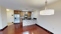 This single-floor residence offers a thoughtfully designed layout centered around a stylish, open-concept kitchen and living area. The kitchen is a standout feature, showcasing warm honey-toned wood cabinetry that provides ample storage, complemented by dark granite countertops that add a touch of elegance. A decorative mosaic tile backsplash adds texture and visual interest, wrapping around both the kitchen walls and the breakfast bar peninsula, which doubles as a casual dining or entertaining space. Stainless steel appliances — including a French door refrigerator with ice and water dispenser, a built-in microwave, and a range — complete the modern kitchen setup. Pendant lighting above the peninsula and a large drum pendant light over the adjoining dining area provide warm, layered illumination throughout the space. Rich, deep-toned hardwood flooring flows seamlessly across the entire living area, grounding the space with warmth and continuity. The open layout between the kitchen and living areas creates an inviting, connected atmosphere ideal for both everyday living and entertaining. The clean white walls and recessed lighting contribute to a bright and airy ambiance throughout this well-maintained residence.