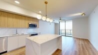 Located along Tampa's iconic Riverwalk Trail in the 33602 zip code, this modern one-bedroom, one-bathroom residence offers a thoughtfully designed open-concept layout with high-end finishes throughout. The combined kitchen and living area serves as the heart of the home, anchored by a striking quartz waterfall island illuminated by a brass and globe pendant light fixture. The kitchen features warm wood cabinetry, a matching quartz backsplash, stainless steel appliances including a French door refrigerator, built-in microwave, and an induction cooktop. Light wood-tone flooring flows seamlessly throughout the main living areas. Floor-to-ceiling sliding glass doors flood the living space with natural light and open to sweeping elevated views of the Tampa skyline and waterfront below, complemented by a ceiling fan overhead. A dedicated hallway leads to the bedroom, which is carpeted and well-proportioned, featuring its own ceiling fan and access to the balcony through large floor-to-ceiling windows. The spacious walk-in closet offers wire shelving for organized storage. The bathroom is appointed with marble-style tile surrounds in the tub-shower combination and warm wood flooring. An in-unit stacked washer and dryer adds everyday convenience to this well-appointed urban residence.
