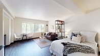 Located along SW Salmon Street in Portland, Oregon, this single-floor studio unit offers a thoughtfully arranged open-concept layout that efficiently combines living, sleeping, and dining areas within one cohesive space. Dark hardwood-style flooring runs throughout, providing a warm contrast to the crisp white walls and ceilings that keep the interior feeling bright and airy.

A generous bank of windows anchors the living area, drawing in abundant natural light and framing lush green tree canopy views that lend a sense of calm to the space. The living zone accommodates a sofa and coffee table alongside a compact dining nook, making good use of the available floor area. An open metal shelving unit serves as a subtle room divider, loosely separating the sleeping area from the lounge space without disrupting the open feel.

The sleeping area comfortably fits a full-size bed and additional seating, with wall art adding personality to the neutral palette. A neatly recessed kitchen alcove houses white upper cabinetry, a gas range, a compact refrigerator, and a sink, providing all essential cooking amenities in an efficient footprint. The bathroom features a full tub-shower combination with clean white fixtures and continues the wood-look flooring found throughout the rest of the unit.