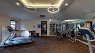 Located at 1700 Seaspray Ct in Houston, Texas, this fitness center is a well-appointed, single-floor facility designed to support a comprehensive workout experience. The space features a modern aesthetic with vaulted ceilings in select areas, complemented by bold circular pendant lighting and recessed cove lighting that create a bright, energizing atmosphere throughout.

The main exercise area is generously proportioned and outfitted with a diverse selection of equipment, including treadmills, elliptical trainers, a stair climber, stationary bikes, and a multi-station strength training cable machine. Free weights and a stability ball round out the available fitness options. Dark, patterned carpet flooring provides both durability and a sleek visual foundation.

A standout feature is the floor-to-ceiling mirrored wall adorned with motivational typographic graphics — featuring words and phrases such as "Aim Higher," "Work Harder," "Power," and "Focus" — adding visual depth and an inspiring ambiance to the workout environment. A wall-mounted flat-screen television is also present for entertainment during workouts.

Natural light filters in through multiple windows, while glass-panel doors provide direct outdoor access, connecting the interior to the surrounding grounds. The facility offers a clean, purposeful design that balances function and style, making it a well-equipped destination for fitness within the community.