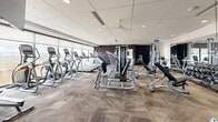 This fitness center in Centennial, Colorado offers a thoughtfully designed, single-floor facility divided into two distinct exercise rooms, providing a comprehensive workout environment for a wide range of fitness goals. The main strength and cardio room features patterned herringbone carpet flooring and is outfitted with an extensive selection of equipment, including cable machines, multi-use weight stations, adjustable benches, a full rack of dumbbells, treadmills, and elliptical trainers. Floor-to-ceiling windows along one wall flood the space with natural light while offering open outdoor views, enhancing the overall workout atmosphere. Ceiling fans and recessed lighting ensure comfort throughout. Large wall mirrors amplify the sense of space and allow users to monitor their form during exercise. A separate multi-purpose studio room features smooth, resilient flooring ideal for group fitness activities and functional training. This room is equipped with spinning bikes, stability balls, a wall-mounted display for guided workouts, foam rollers, resistance accessories, and additional free weights. Textured accent wallcovering adds a modern design touch, while a large mirror and natural light from oversized windows create an open, energizing environment. Together, the two rooms offer a versatile and well-appointed fitness experience.