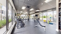 Located at 216 First St in West Palm Beach, Florida, this single-floor fitness center is designed to provide a comprehensive workout experience in a bright, modern environment. The space is defined by expansive floor-to-ceiling windows along multiple walls, flooding the interior with natural light and offering open views of the surrounding landscaped grounds and outdoor amenities.

The main workout floor features durable rubberized flooring and is outfitted with a wide array of cardio equipment, including treadmills, ellipticals, and stationary bikes from Matrix. A dedicated strength training zone includes multi-station cable machines, adjustable weight benches, and a full rack of dumbbells neatly arranged on tiered stands alongside built-in wood shelving. Floor-to-ceiling mirrors along one wall visually expand the space and support proper form during exercise.

A functional training area offers stability balls, resistance bands, yoga mats, and suspension training equipment, catering to a variety of workout styles. Motivational typography wraps the walls throughout, featuring phrases such as "Goal Strong," "Repetitions," "Motivation," and "Workout Energy Fitness," contributing to an energizing atmosphere. The drop ceiling with recessed panel lighting ensures consistent illumination across the entire facility, complementing the abundant daylight for a vibrant, performance-focused environment.