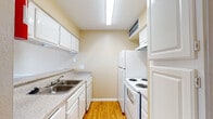 This single-story space is designed with a functional, cohesive layout that flows naturally between its key areas. The galley-style kitchen is efficiently organized, featuring white cabinetry above and below granite-look countertops, a double stainless steel sink, a white electric coil range, and a refrigerator. Warm honey-toned hardwood-style flooring runs the length of the kitchen, complementing the soft cream-colored walls and overhead fluorescent lighting. A dishwasher is integrated beneath the counter, maximizing utility within the compact corridor layout.

The living area is finished with plush wall-to-wall carpeting in a neutral beige tone and is highlighted by a white-surround fireplace, serving as a natural focal point. Crown molding lines the ceiling, adding a refined architectural detail, while a window fitted with horizontal blinds allows natural light to filter into the space. An adjacent area, likely a dining space, features the same carpeting and crown molding, with a pendant light fixture overhead and a chair rail detail along the walls.

The bathroom includes a single vanity with a granite-look countertop, an undermount basin sink, a framed mirror, and standard fixtures, maintaining the warm neutral palette found throughout the unit.