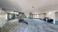 Located at 2301 S Valley View Blvd in Las Vegas, NV, this fitness facility spans a generous single-floor footprint and is divided into two dedicated exercise rooms, each thoughtfully designed to support a variety of training styles. The main exercise area features patterned carpet tile flooring throughout, multiple ceiling fans for air circulation, and recessed lighting that keeps the space bright and functional. This room is equipped with a multi-station cable machine, weight benches, a rack system, cardio machines including treadmills, stationary bikes, an elliptical, and a rowing machine. Motivational wall murals with bold typography — including phrases like "Success Starts with Discipline" and "Commit to Fitness" — are printed directly onto textured, concrete-style accent walls, contributing to an energizing atmosphere. A second exercise room offers a more versatile functional training environment, highlighted by a colorful bouldering/climbing wall, exercise mats, a mini trampoline, plyo boxes, and a dumbbell rack. Inspirational quotes adorn the walls here as well, reinforcing the motivational theme. Both rooms benefit from natural light through street-facing windows and glass entry doors, providing views to the exterior. The overall design is clean, modern, and purposefully organized to accommodate diverse fitness goals.