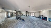 Located at 2301 S Valley View Blvd in Las Vegas, NV, this fitness facility spans a generous single-floor footprint and is divided into two dedicated exercise rooms, each thoughtfully designed to support a variety of training styles. The main exercise area features patterned carpet tile flooring throughout, multiple ceiling fans for air circulation, and recessed lighting that keeps the space bright and functional. This room is equipped with a multi-station cable machine, weight benches, a rack system, cardio machines including treadmills, stationary bikes, an elliptical, and a rowing machine. Motivational wall murals with bold typography — including phrases like "Success Starts with Discipline" and "Commit to Fitness" — are printed directly onto textured, concrete-style accent walls, contributing to an energizing atmosphere. A second exercise room offers a more versatile functional training environment, highlighted by a colorful bouldering/climbing wall, exercise mats, a mini trampoline, plyo boxes, and a dumbbell rack. Inspirational quotes adorn the walls here as well, reinforcing the motivational theme. Both rooms benefit from natural light through street-facing windows and glass entry doors, providing views to the exterior. The overall design is clean, modern, and purposefully organized to accommodate diverse fitness goals.