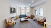 Located at 51 Queensberry St in Boston's Fenway neighborhood, this single-floor space blends historic architectural character with contemporary design. The main lounge area is anchored by a charming bay window arrangement featuring original-style window frames with decorative stained glass transoms, allowing generous natural light to fill the room. The seating area is thoughtfully furnished with a tufted teal sofa, accent chairs, a wood coffee table, and a wall-mounted flat-screen TV, creating a comfortable and functional gathering environment. Patterned area rugs define the lounge zone over light wood-style flooring that extends throughout the space. A connecting corridor, adorned with vibrant abstract artwork, leads toward a glass-panel entry door, lending the space an open and airy feel. The refreshment area features a wall-mounted mini-split HVAC unit, a white cabinetry station stocked with beverages and snacks, and additional appliances, making it well-equipped for daily use. Recessed lighting throughout ensures consistent illumination across all areas. The overall ambiance is clean, modern, and inviting, with warm tones and curated furnishings that balance both function and comfort — well-suited for use as a communal lounge, waiting area, or shared amenity space.