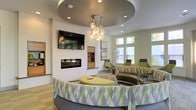 This single-floor communal lounge is designed with a contemporary aesthetic that balances comfort and sophistication. The centerpiece of the space is a curved, circular sofa upholstered in a bold geometric pattern of chartreuse, gray, and white, complemented by a matching rounded ottoman. Above, a recessed circular tray ceiling frames a cluster-style pendant chandelier with exposed Edison-style globe bulbs, adding a warm, artistic focal point.

A wall-mounted flat-screen television is positioned above a sleek, linear gas fireplace, flanked by built-in shelving that provides both functional storage and display space. The surrounding walls are finished in a clean white, enhancing the brightness of the room.

Natural light filters in through multiple windows along the far wall, offering views of greenery outside. The seating area near the windows features modern accent chairs in a coordinating green tone, along with a sculptural arc floor lamp. Light wood-toned flooring runs throughout, grounding the space with warmth. The overall ambiance is polished, inviting, and well-suited for social gatherings or relaxed conversation.