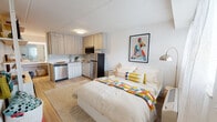 This well-appointed studio offers a thoughtfully designed single-level open-plan layout that maximizes every square foot. The living and sleeping area is bright and welcoming, featuring light wood-tone flooring, crisp white walls, and a large window dressed with sheer curtains that allow natural light to fill the space. A comfortable full-sized bed serves as the room's focal point, complemented by open shelving that provides both storage and display space, along with a mounted flat-screen television.

The kitchen is seamlessly integrated into the living space and is outfitted with light wood cabinetry, a granite-style countertop, a subway tile backsplash, and a full suite of stainless steel appliances including a refrigerator, dishwasher, built-in microwave, and a smooth-top range. The clean, contemporary finishes give the kitchen a modern and functional aesthetic.

The bathroom features a walk-in shower with white tile surround, a shower curtain, and a toilet, accented by colorful framed artwork that adds a lively touch. Throughout the studio, pops of color from carefully chosen art pieces and textiles create a cheerful, curated ambiance that balances practicality with personality.