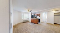 This well-organized single-floor unit offers a functional and efficient layout across a compact footprint. The open-concept living and kitchen area forms the heart of the space, where soft carpet in the living zone transitions to tile flooring in the kitchen. A central peninsula with granite countertops serves as both a workspace and a divider between the two areas, topped with a ceiling fan that provides additional air circulation. The kitchen is well-equipped with warm medium-toned wood cabinetry, granite countertops, a full suite of black appliances including a refrigerator, electric range, and over-the-range microwave, along with a stainless steel sink beneath a window that invites natural light. Multiple windows throughout the unit ensure consistent natural light across all rooms. The bedroom is a comfortable, carpeted space featuring two windows with blinds, a ceiling fan with light fixture, and a wall-mounted HVAC unit. The bathroom is finished with large-format marble-style tiles covering both the floors and tub surround, complemented by a white vanity and a glass-block window above the tub, adding brightness while maintaining privacy.